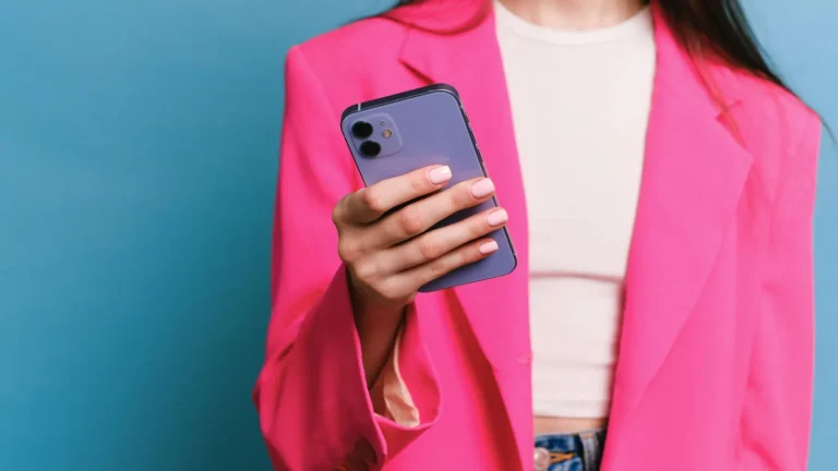 Woman in a bright pink blazer holding a current smartphone against a blue wall