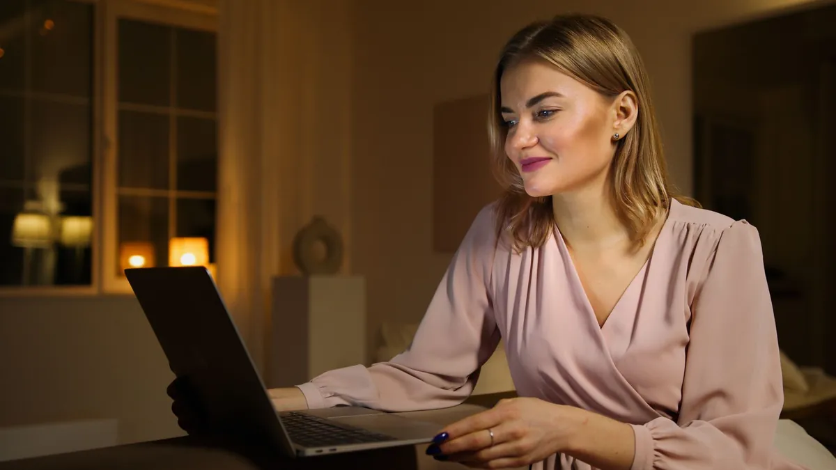 Smiling woman in a blush blouse working on a laptop in a softly lit room in the evening