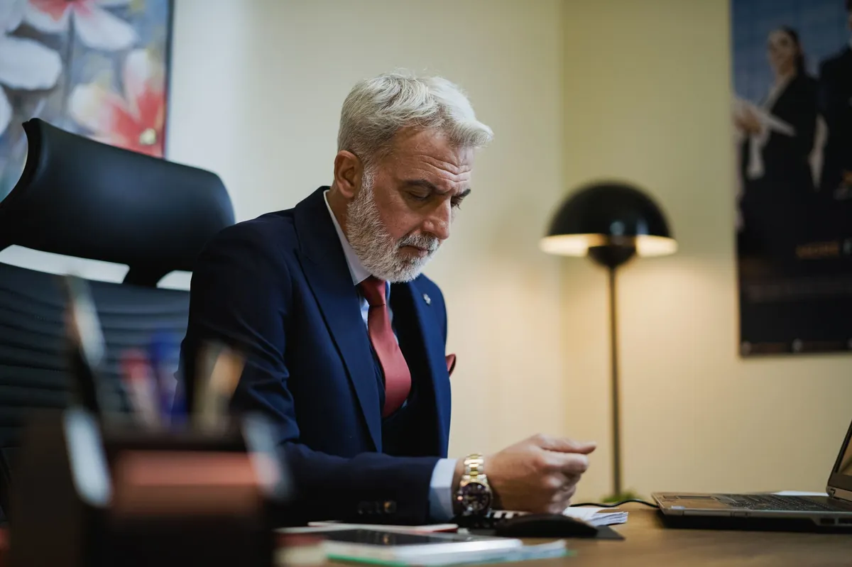 Silver-haired executive reviewing papers at a desk beside a floor lamp